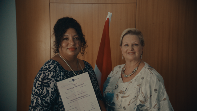 Eine Frau zeigt vor der Kamera ein Dokument mit der Aufschrift „Bescheid“.