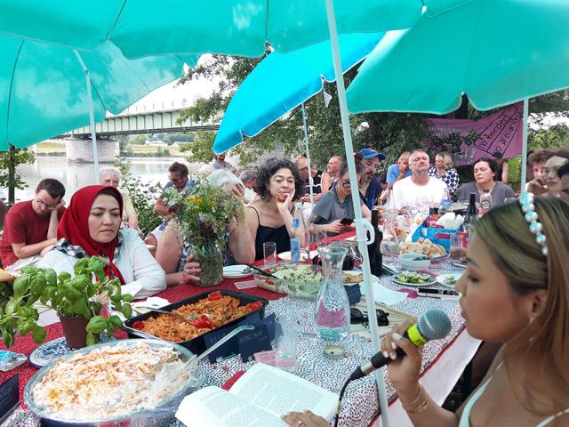 Unter einem Sommerzelt sitzen Menschen und hören einer Person zu, die ins Mikrofon spricht. Auf dem Tisch stehen Teller mit Essen.