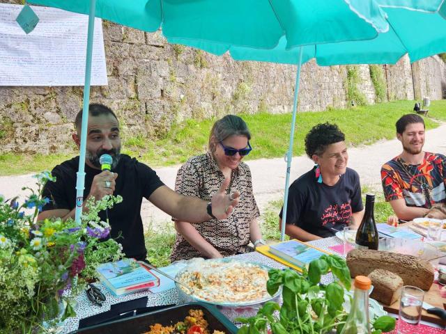 Vier Personen sitzen im Freien unter einem Sommerzelt, während eine von ihnen ins Mikrofon spricht. 