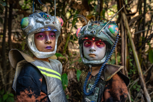 Zwei Kinder blicken in die Ferne, gekleidet in Stofffetzen und mit rotem Make-up rund um die Augen.